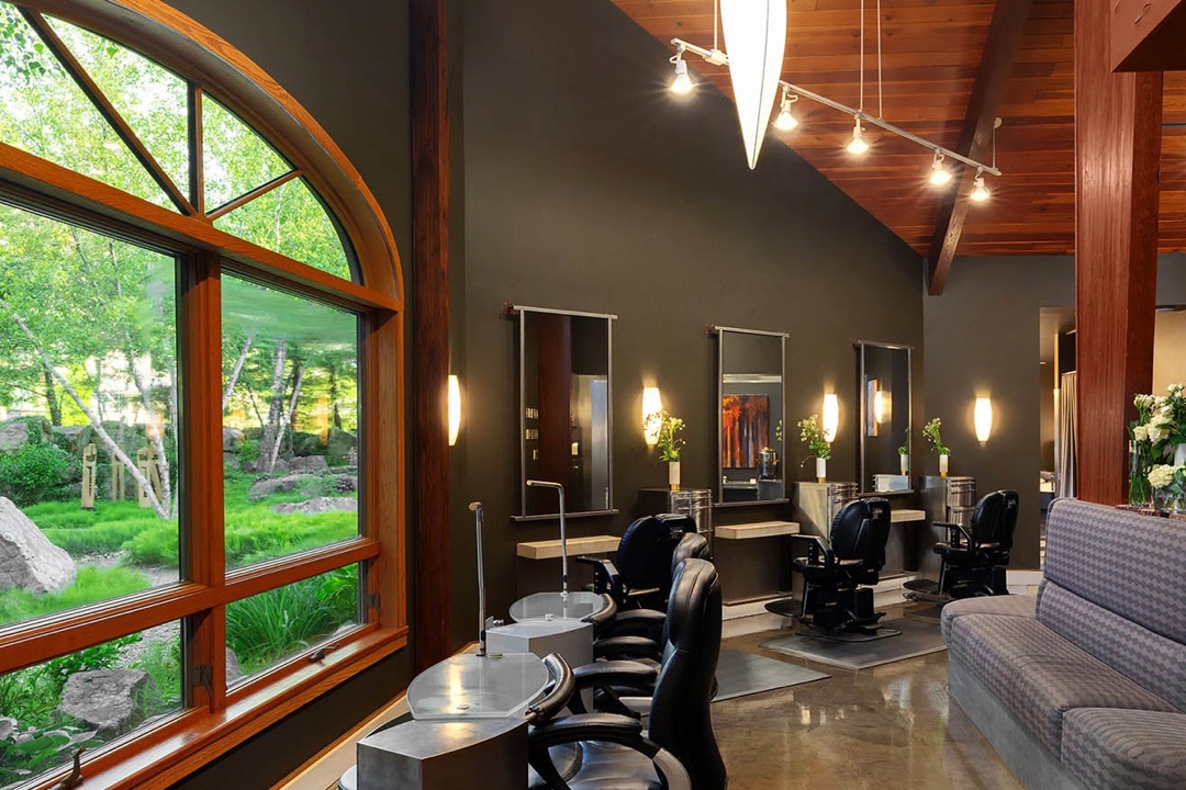 the interior shot of a beauty salon. there are plush black leather chairs in front of mirrors and nail painting tables. There are windows with a view of nature