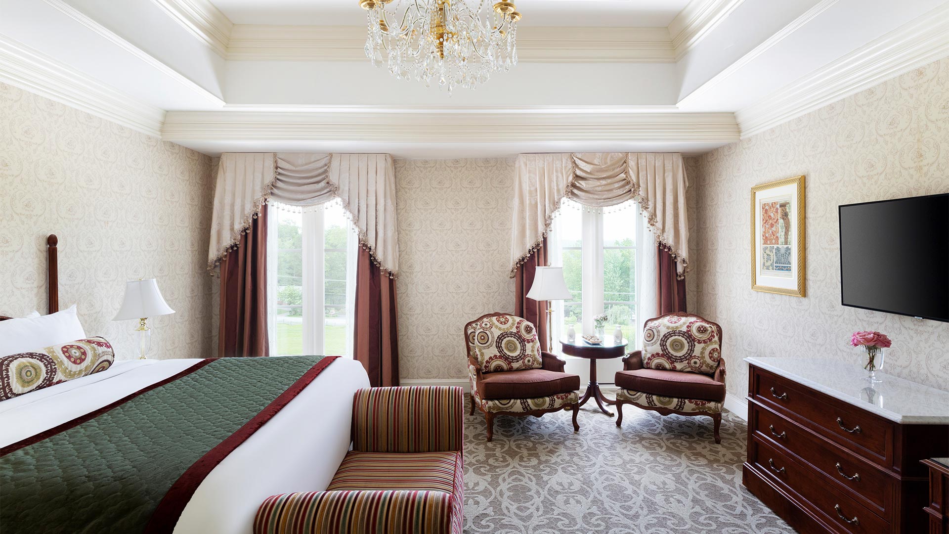 interior shot of The Chateau's king accommodation. There is a king bed with white and green linens. The room also includes views of the resort grounds, a small sitting area and a tv with a dresser.