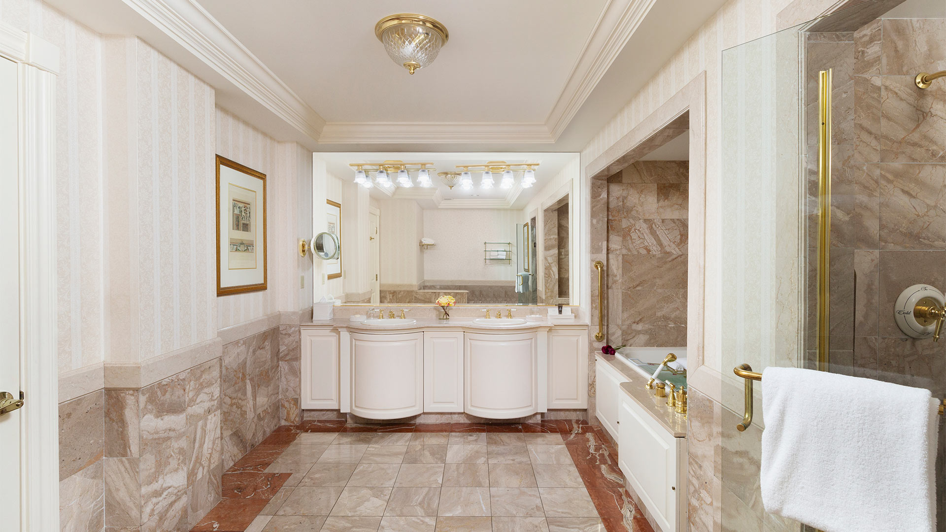 interior shot of a bathroom. the colors are a neutral beige. There is an elegant double sink with a large mirror and a shower and bath combination to the right of the sinks.