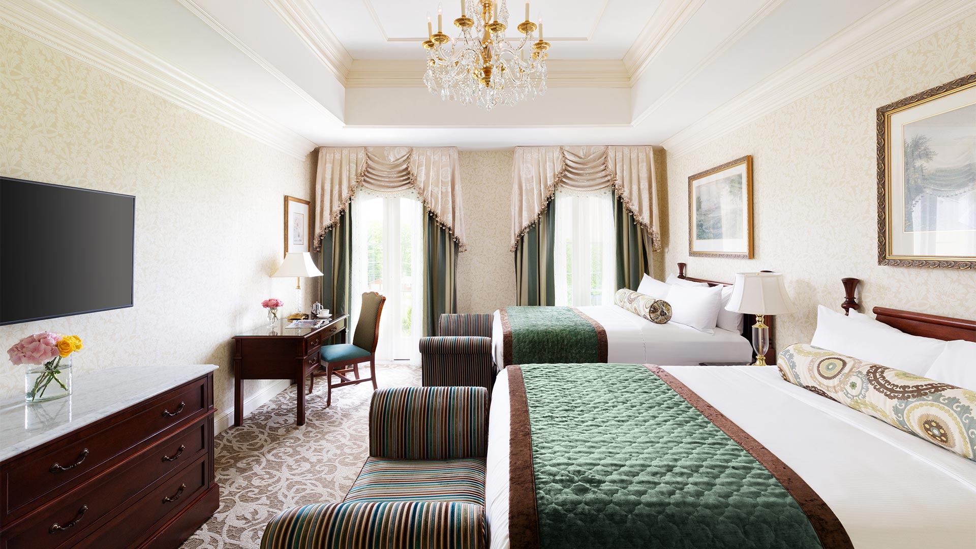 interior shot of the bedroom for The Chateau's Double Suite. There are two queen beds with white and green linens and a sitting bench at the foot of each. There is a desk and tv across from the beds.