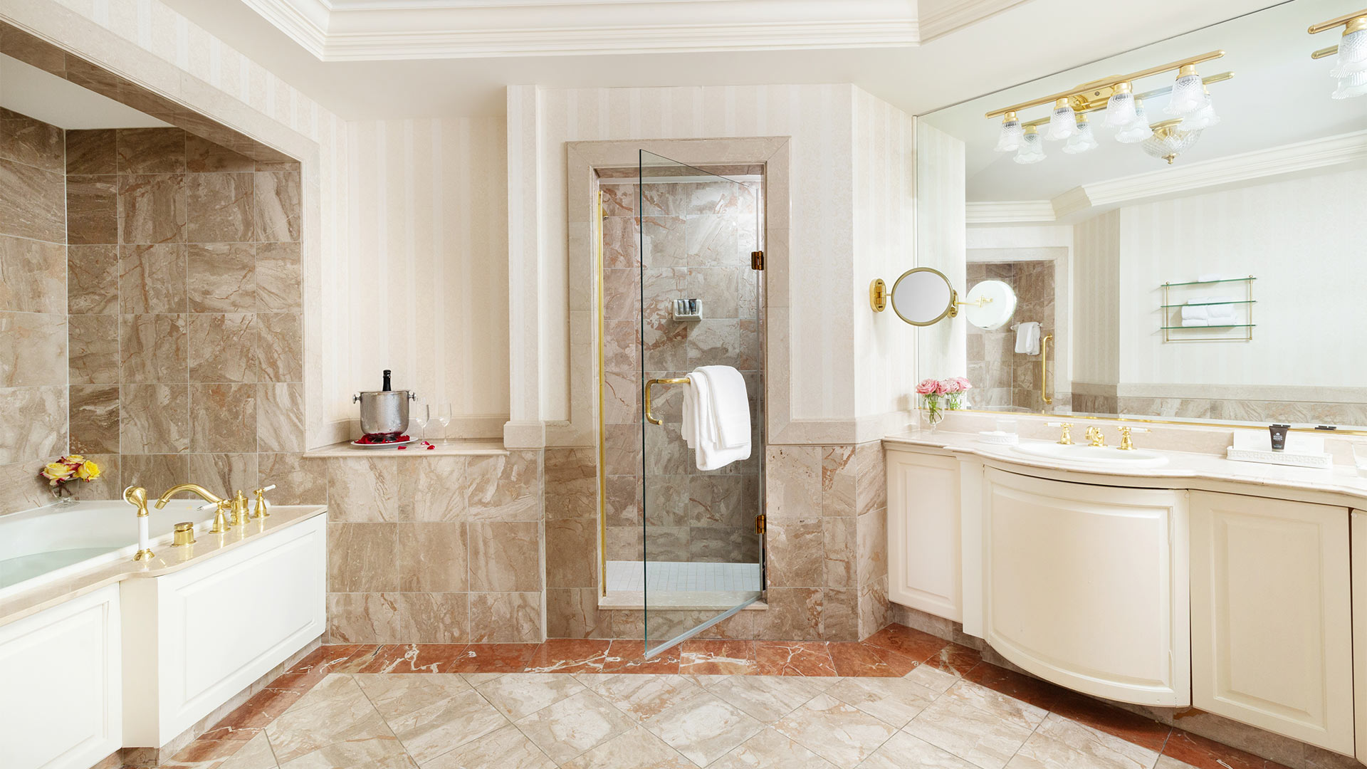 interior shot of the suite's bathroom. The bathroom has neutral beige tiling. There is a large sink and mirror across from a whirlpool bathtub. In between the sink and bathtub is a walk in shower.