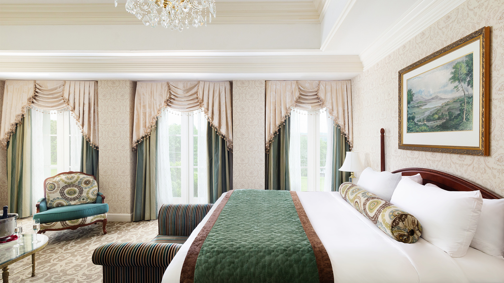 interior shot of The Chateau's Junior King Suite bedroom. There is a king bed with green and white linens with a sitting bench on the end of the bed. There is a sitting area and three large windows overlooking the resort grounds.