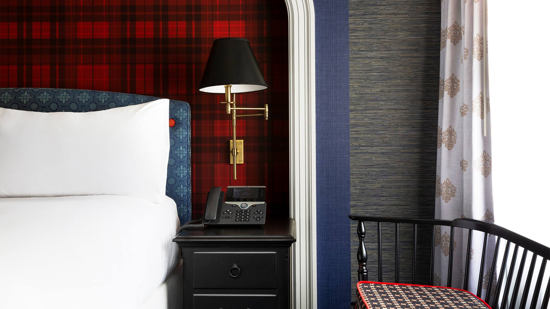 detail shot of one of the townhomes' bedrooms. The bed has crisp white bedding, the wall behind the bed is a cozy buffalo plaid pattern. There is a nightstand, a lamp and a chair to the right of the bed.
