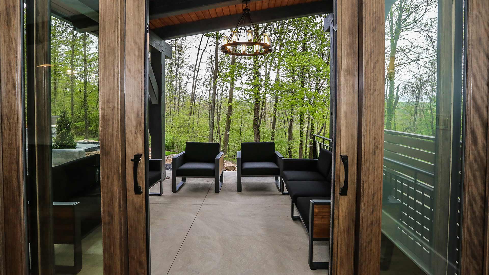 exterior shot of the back deck and yard. There are chairs and a couch on the patio. There is a view of the woods in the background.