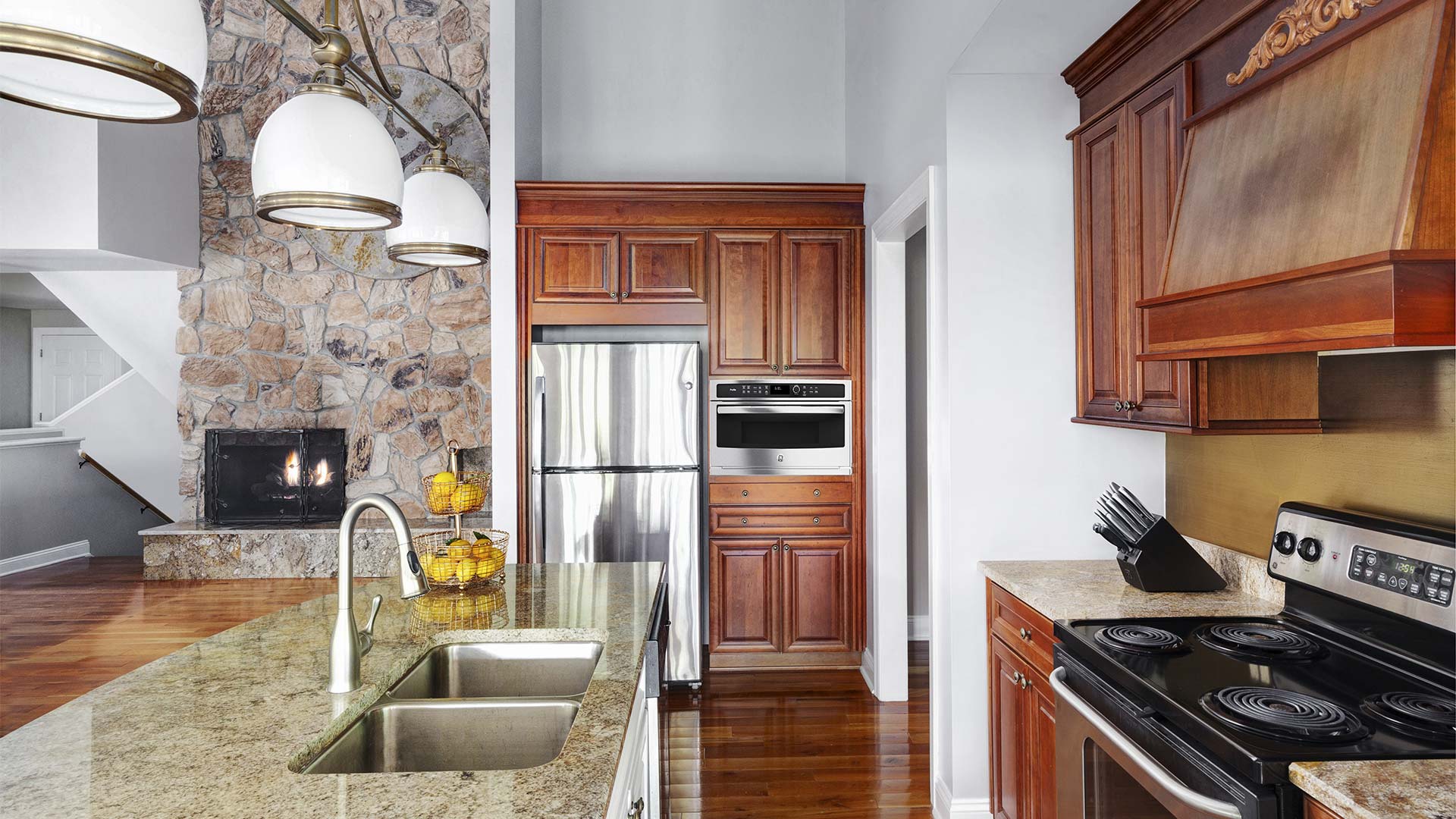interior shot of garshak's kitchen. There is a center island with two sinks. There is a full fridge and microwave in the background surrounded by dark wood cabinets. Across from the sink is the stovetop and more counter space and overhead cabinets.