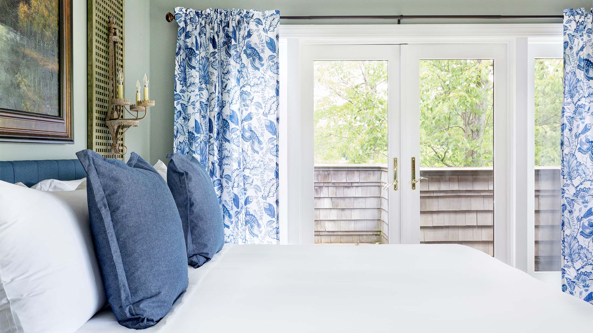 detail shot of a bedroom. There is a bed with white linens and blue throw pillows. There are glass doors that lead out to a deck area. There are blue floral curtains.