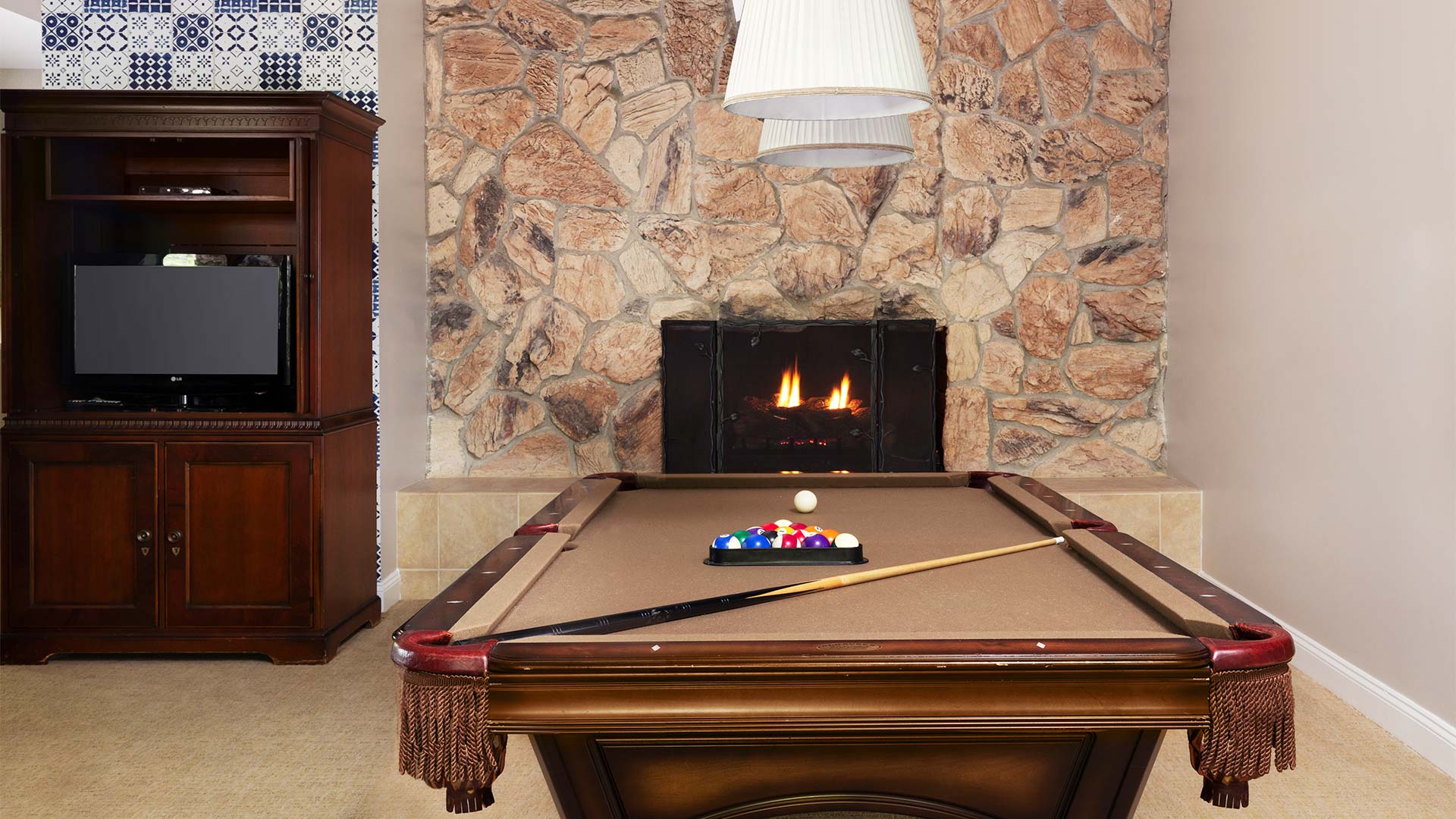 interior shot of a game room. There is a pool table with the balls set up and a pole resting on the table. There is a fireplace in the background.