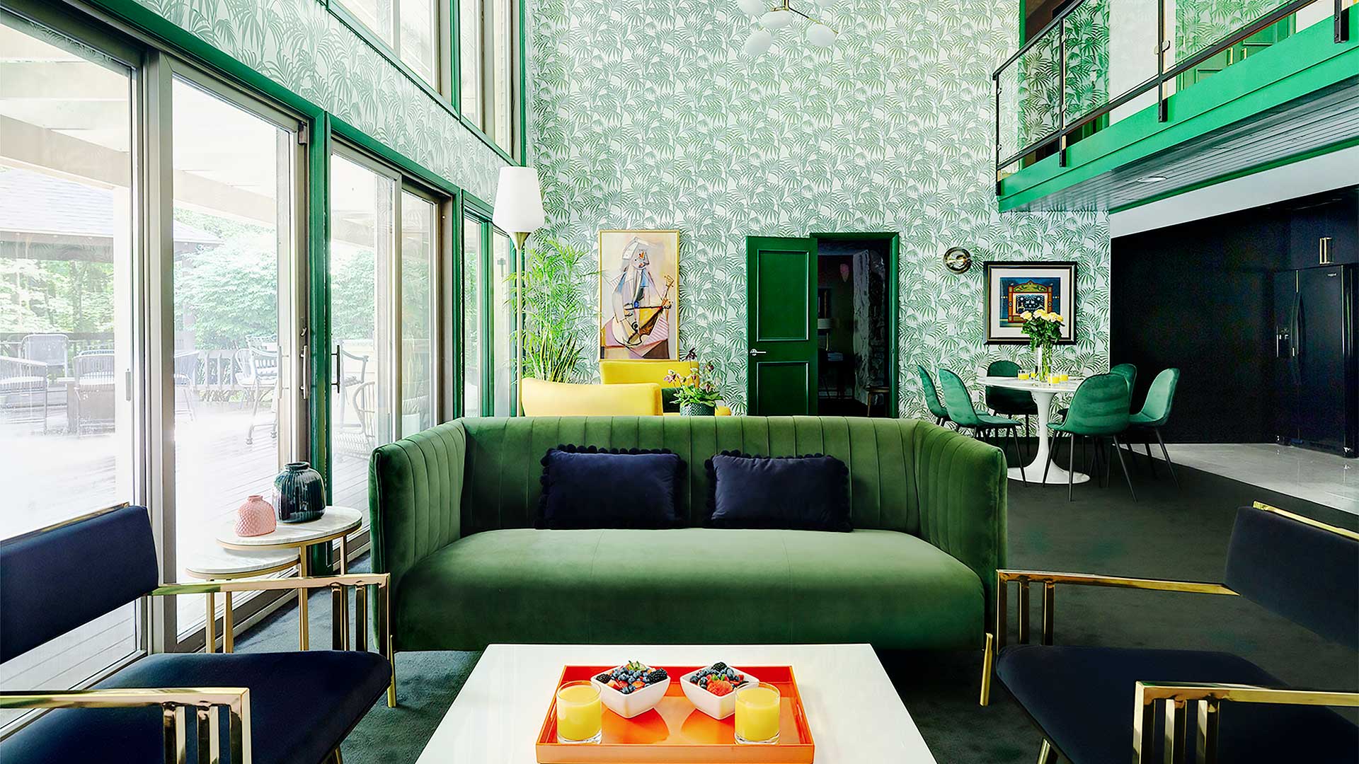an interior shot of grouse glen's living area. The space has a stunning emerald green theme. The couch is velvet and there is patterned wallpaper in the background. Art dots the room bringing in accents of orange, red and yellow