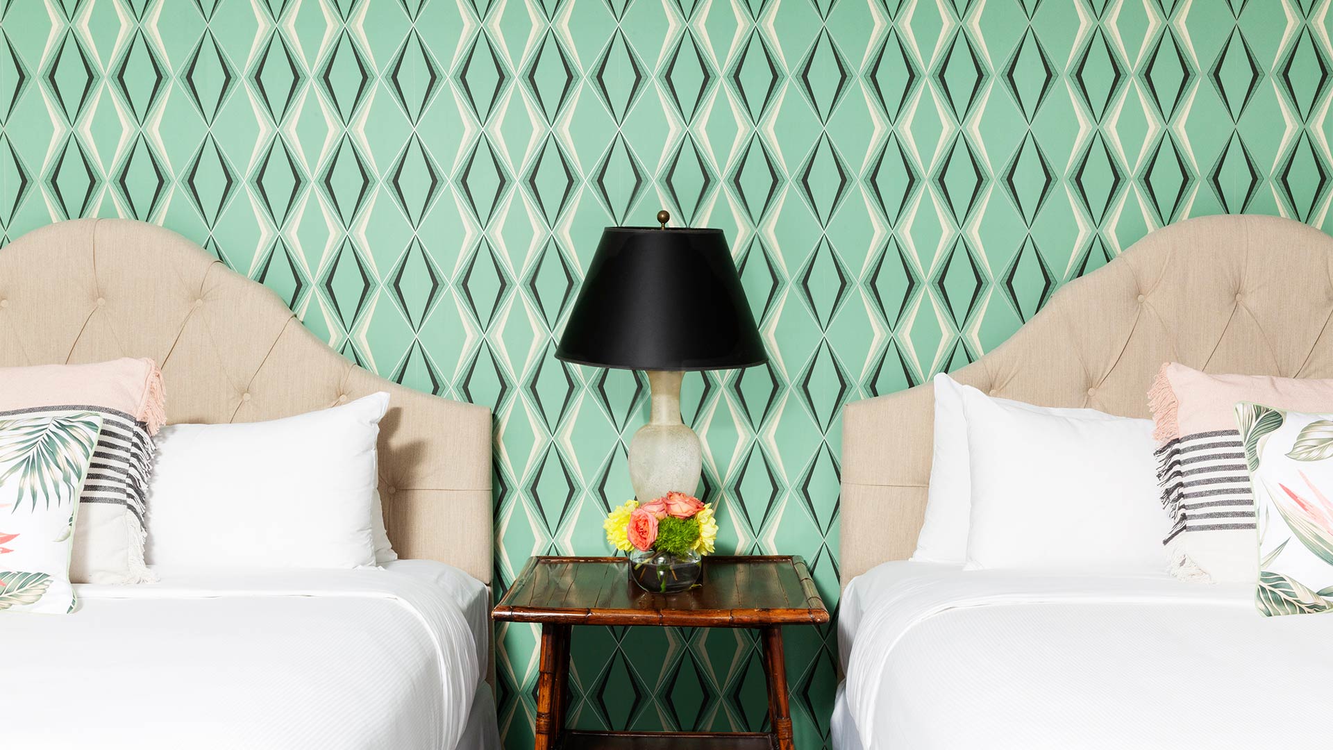 a detail shot of a bedroom with two twin beds. They are separated by a beside table with a lamp and flowers on top of it. There is a retro-inspired green patterned wallpaper behind the two beds.