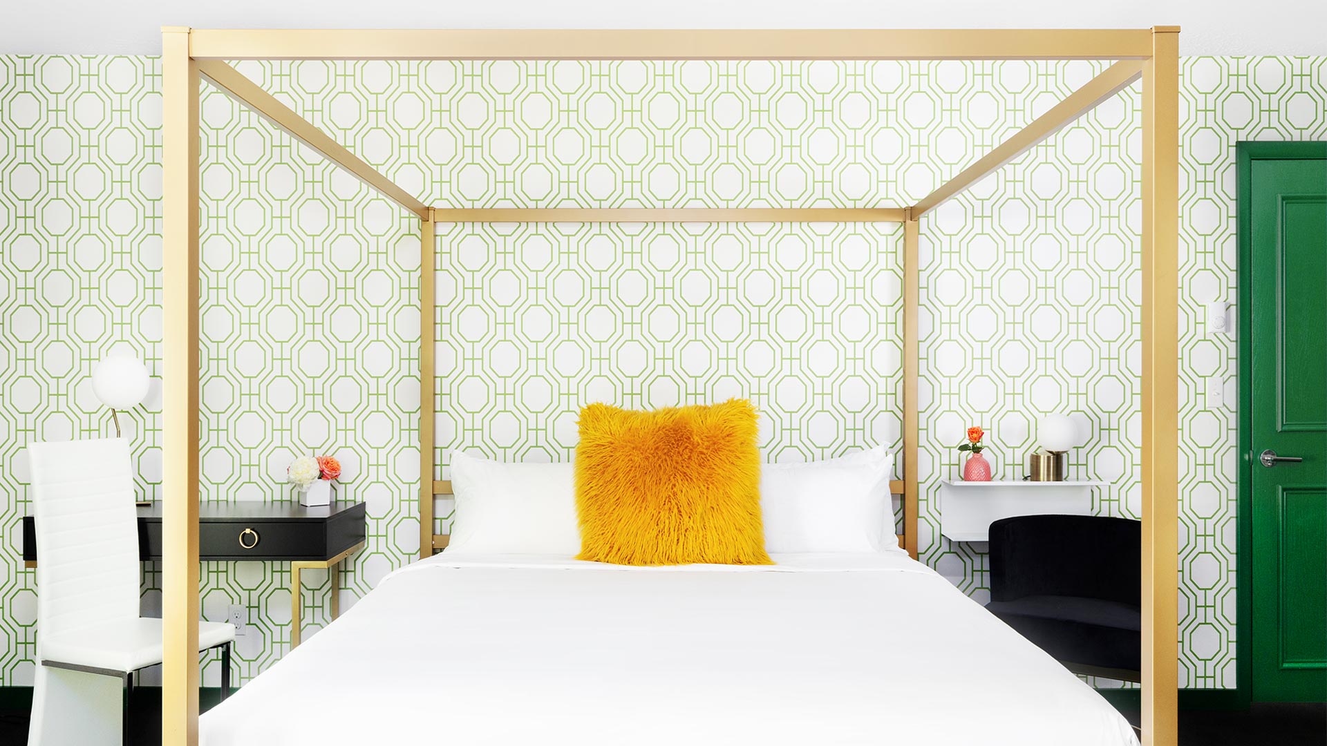 detail shot of a bedroom with a canopy bed. the bed has white linens with a yellow fluffy accent pillow. There are tables on either side of the bed. There is green retro-inspired wallpaper behind the bed
