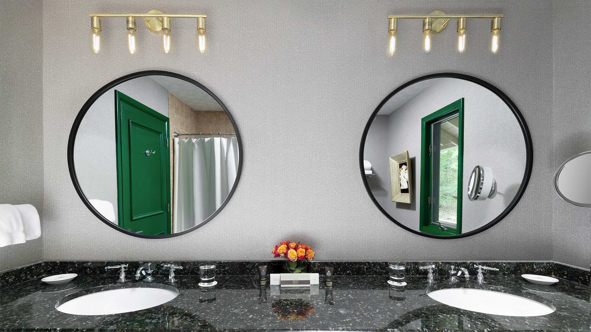 detail shot of a bathroom. There are two sinks with a sleek dark grey countertop. There are circular mirrors above each sink.