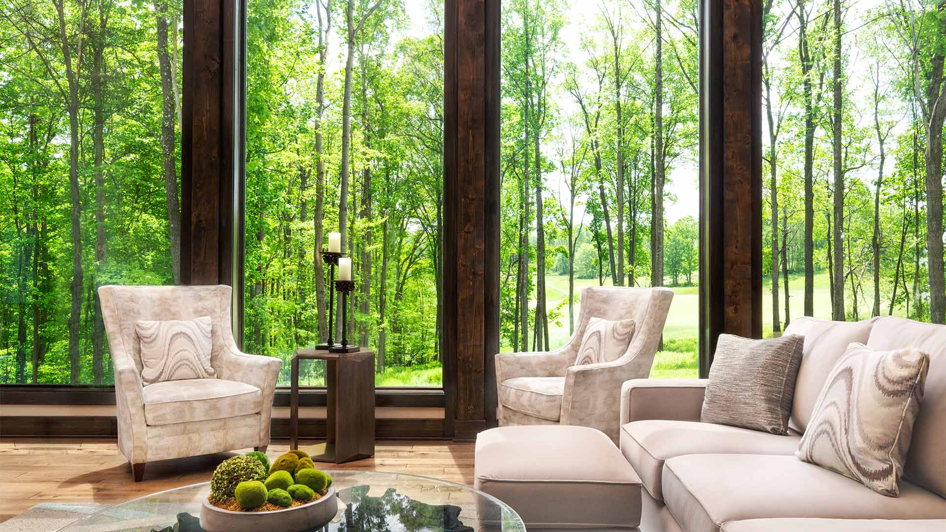 interior shot of the dogwood sitting area. There are two cream colored chairs and a matching couch around a coffee table. There are floor to ceiling windows give a view of the forest outside
