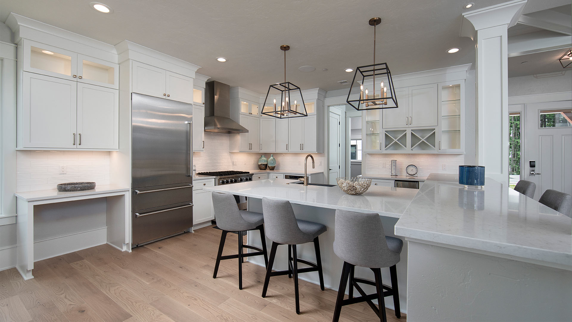 interior shot of a kitchen. The countertops and cabinets are a sleek white. There are gray chairs lining the kitchen island. There are stainless steel appliances.