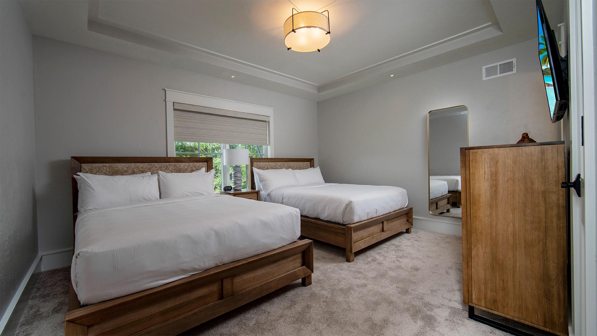 interior shot of a bedroom with two queen beds. Each bed has all white linens and wooden headboard. There is a dresser with a tv mounted overhead across from the beds