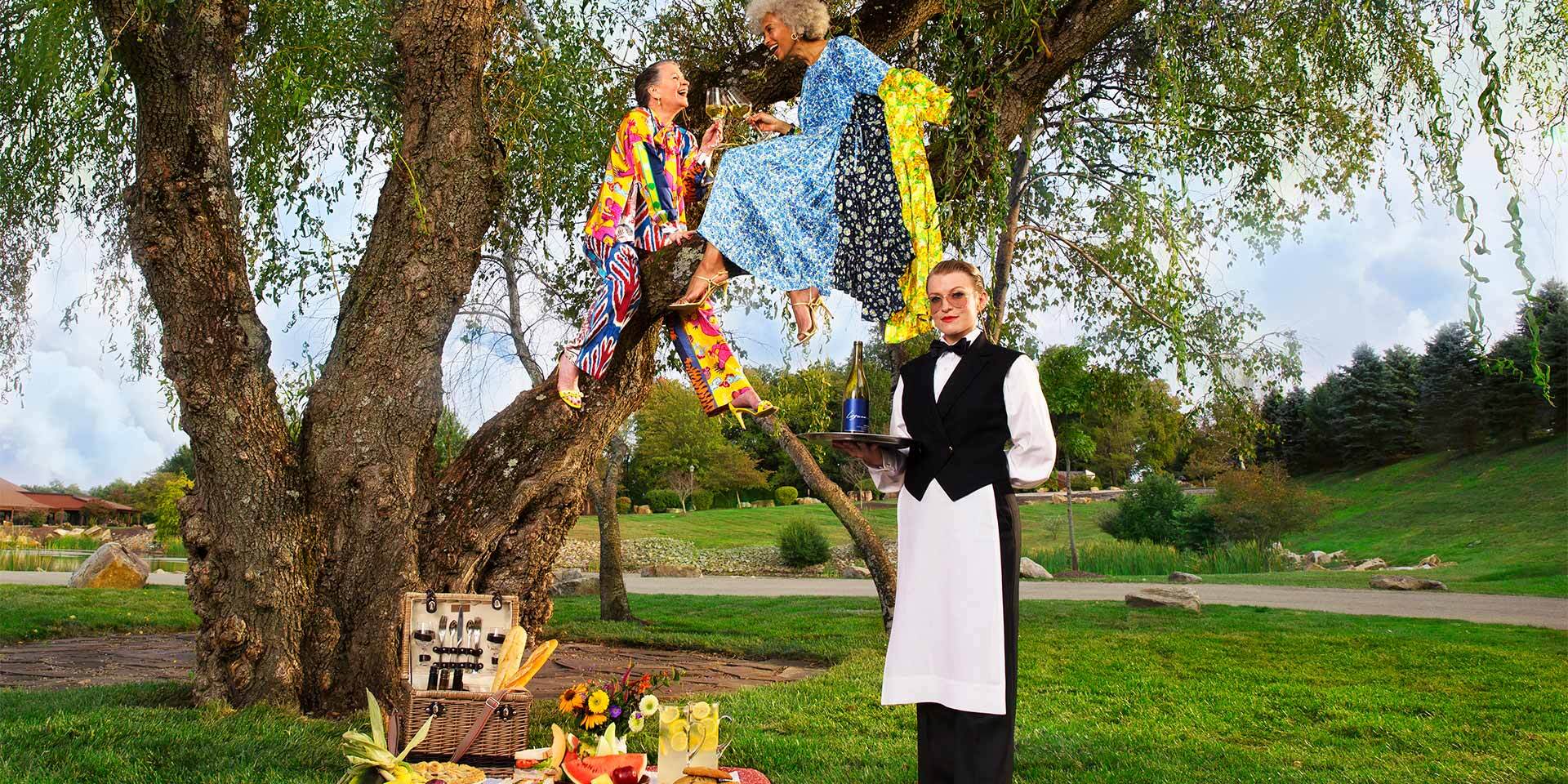 Two older women sit in a tree and enjoy a picnic on the ground catered to them by a woman standing in front of them with champagne in hand.