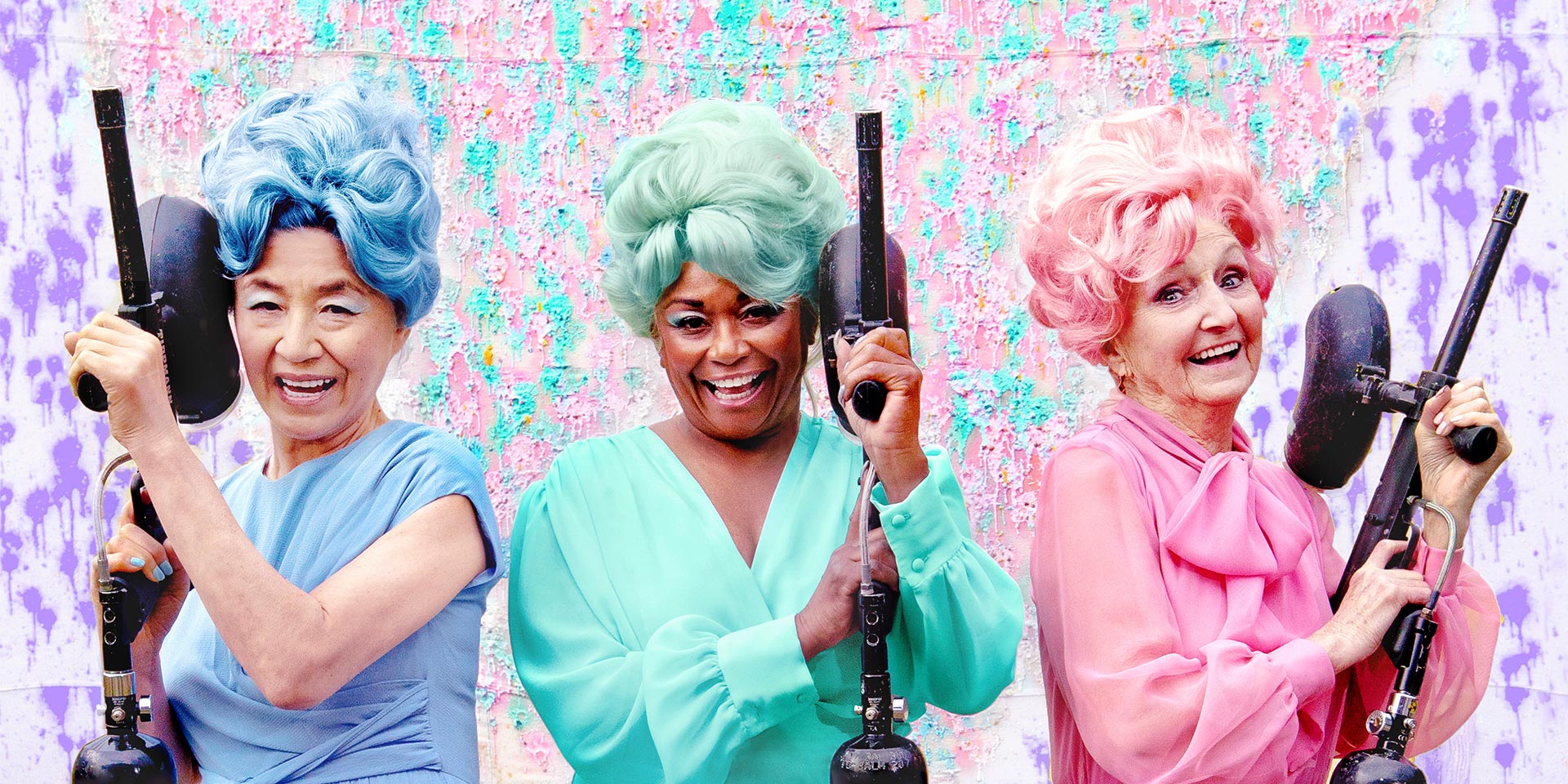 Three older women wearing monochrome outfits with wigs, one blue, one green, one pink, in front of a colorful backdrop