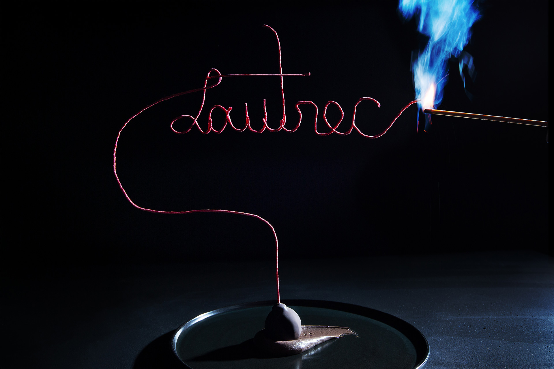 A fancy wick that spells "Lautrec" in cursive being lit by a match