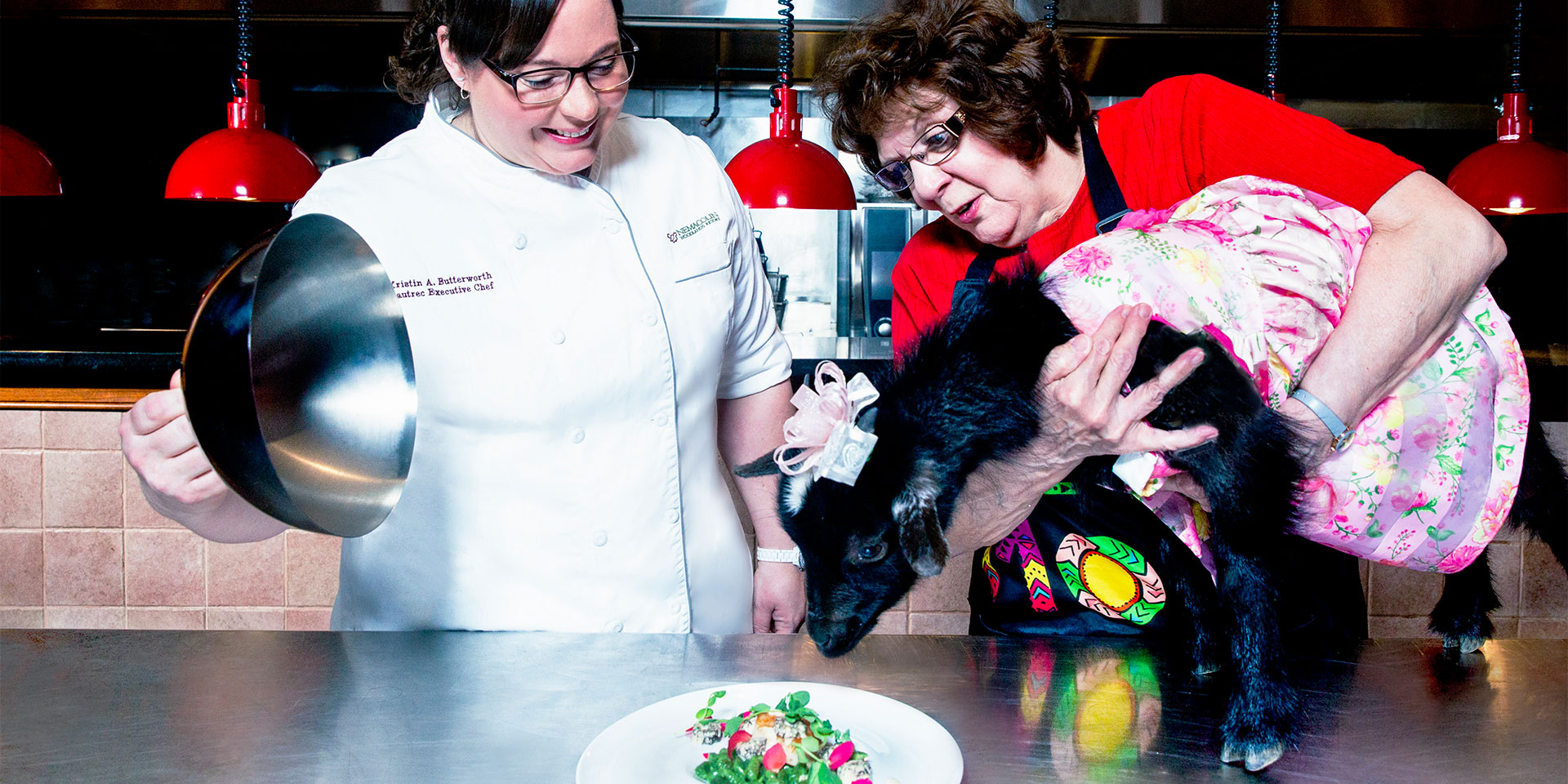 a chef revealing a plate and a woman holding a goat in a dress up to the plate
